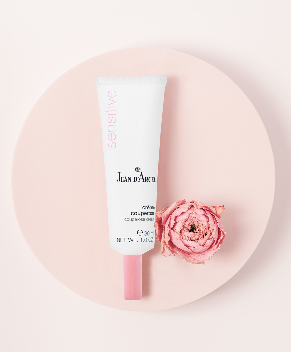 Pflege-Tube liegt auf einem runden Podest, daneben eine rosa Blüte.